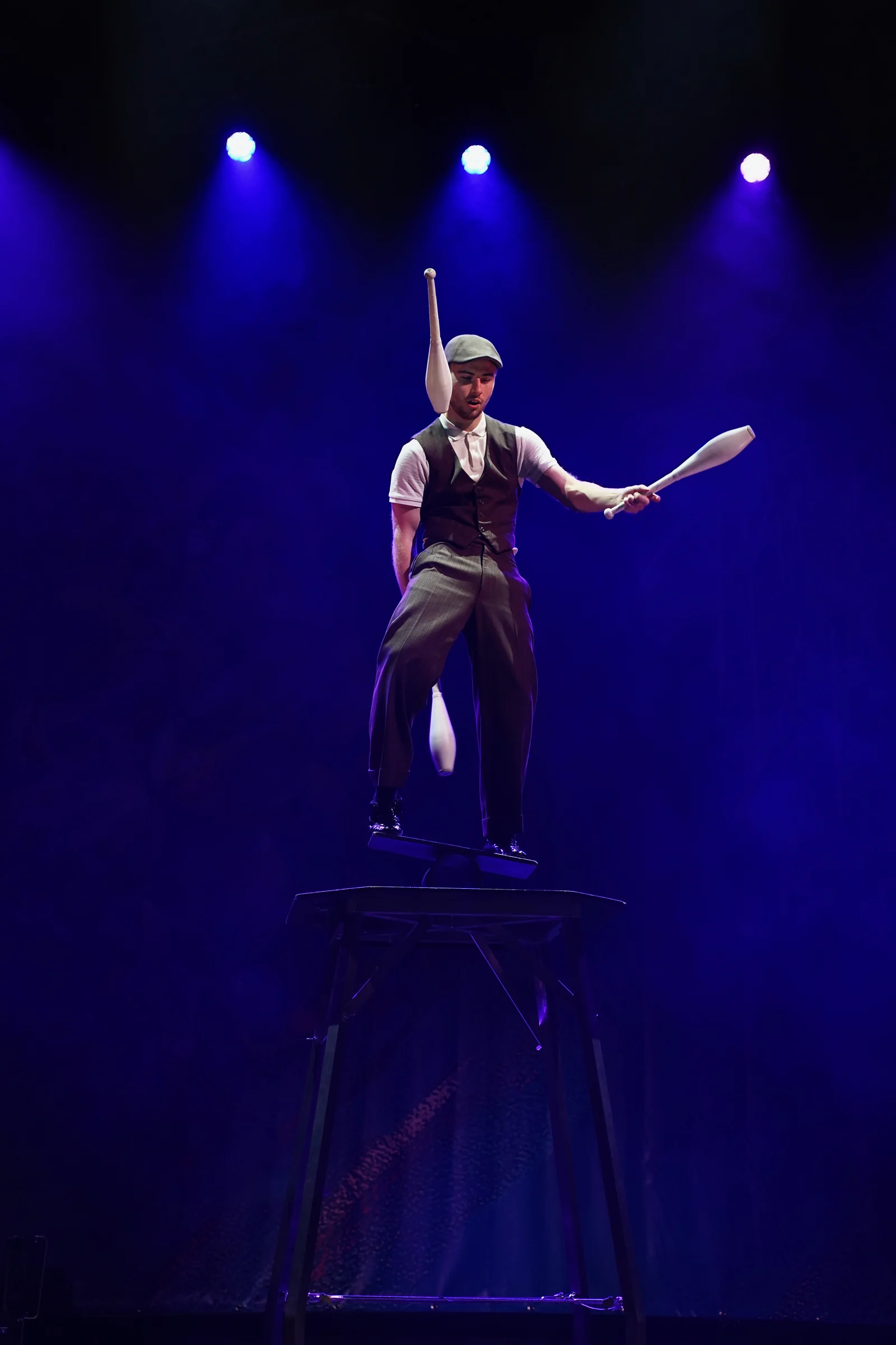 Misha juggling clubs on a rola bola board under blue stage lights